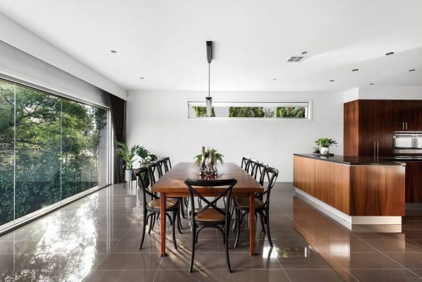 Prestige New Home with brown tiled flooring, a large will length window, brown kitchen bench with black top and wooden dining table and chairs.