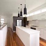 Custom built home with white modern kitchen with centre bench, 3 black hanging lights above bench, polished wooden floor boards and staircase on left.