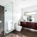 Dark wood bathroom vanity, shower with full length glass partition, white toilet in custom built home
